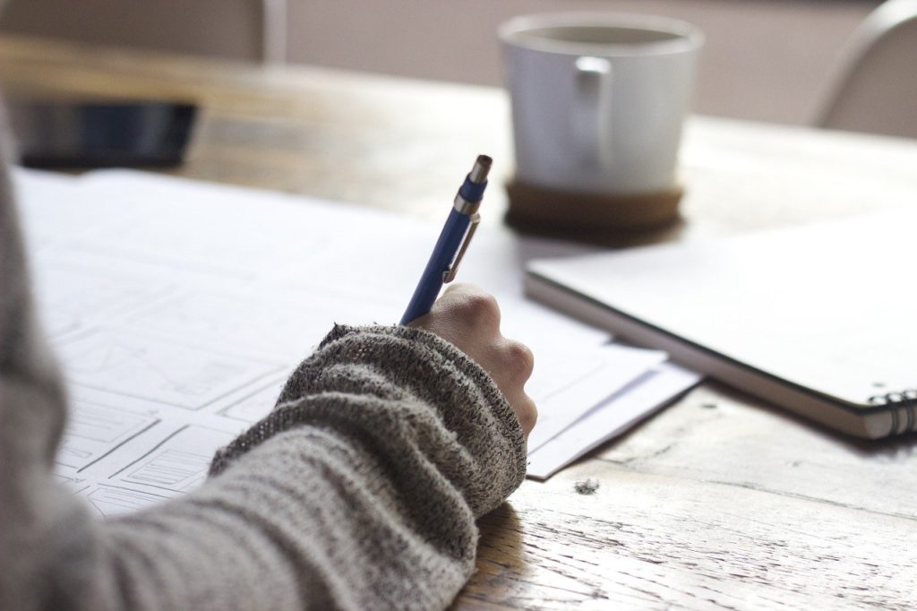 Forearm wearing grey jumper holding a pen, working on documents.  White coffee cup in the background on a timber table.